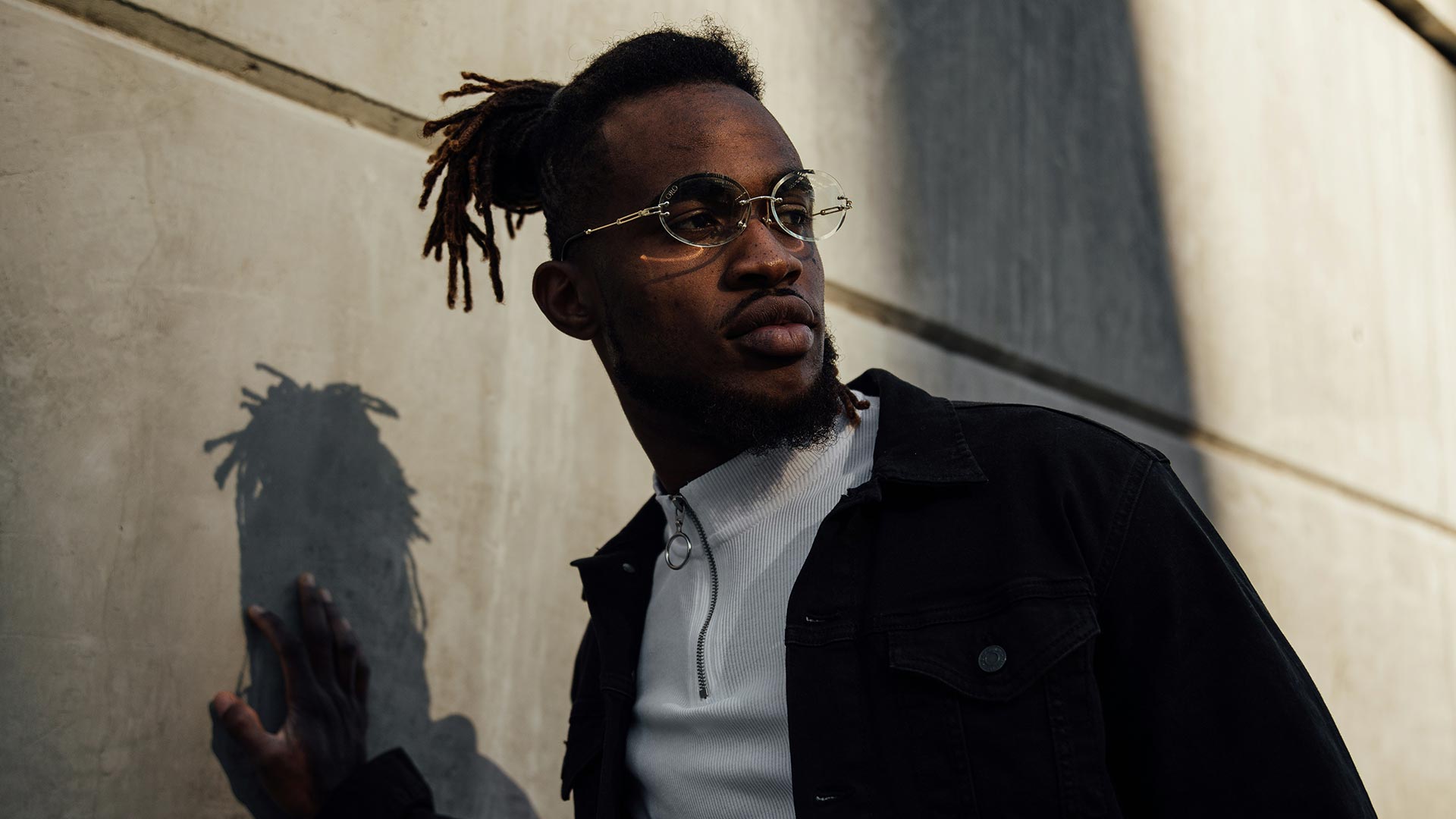 Selbstbewusster junger Mann mit Dreads trägt eine runde Metallbrille mit kleinen Gläsern und lehnt an einer Betonwand – neue Brillenmodelle für Herren bei Seitz Optik Limburg