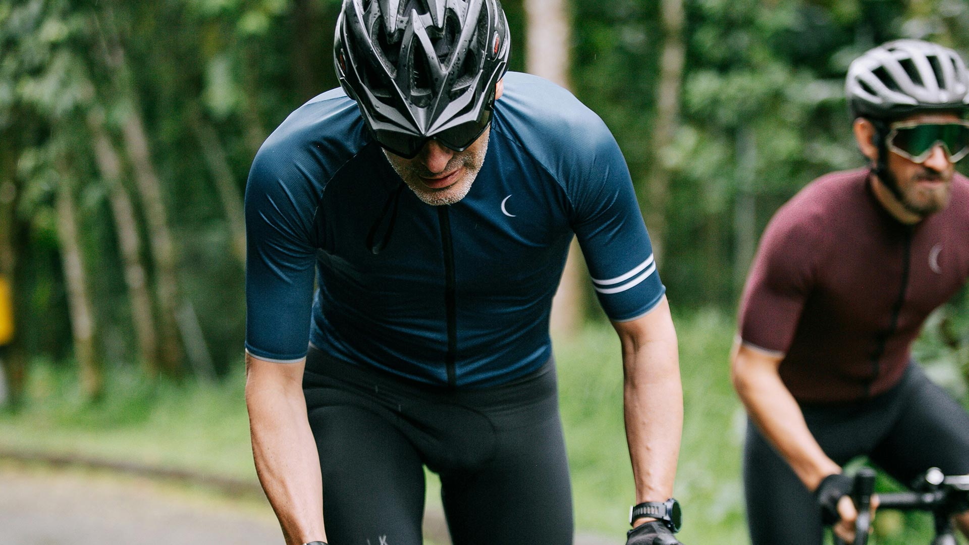 Zwei Radsportler mit Sportbrillen und Helmen bei einem Rennen auf einer Straße umgeben von Wald – robuste Sportbrillen für aktive Menschen bei Seitz Optik Limburg